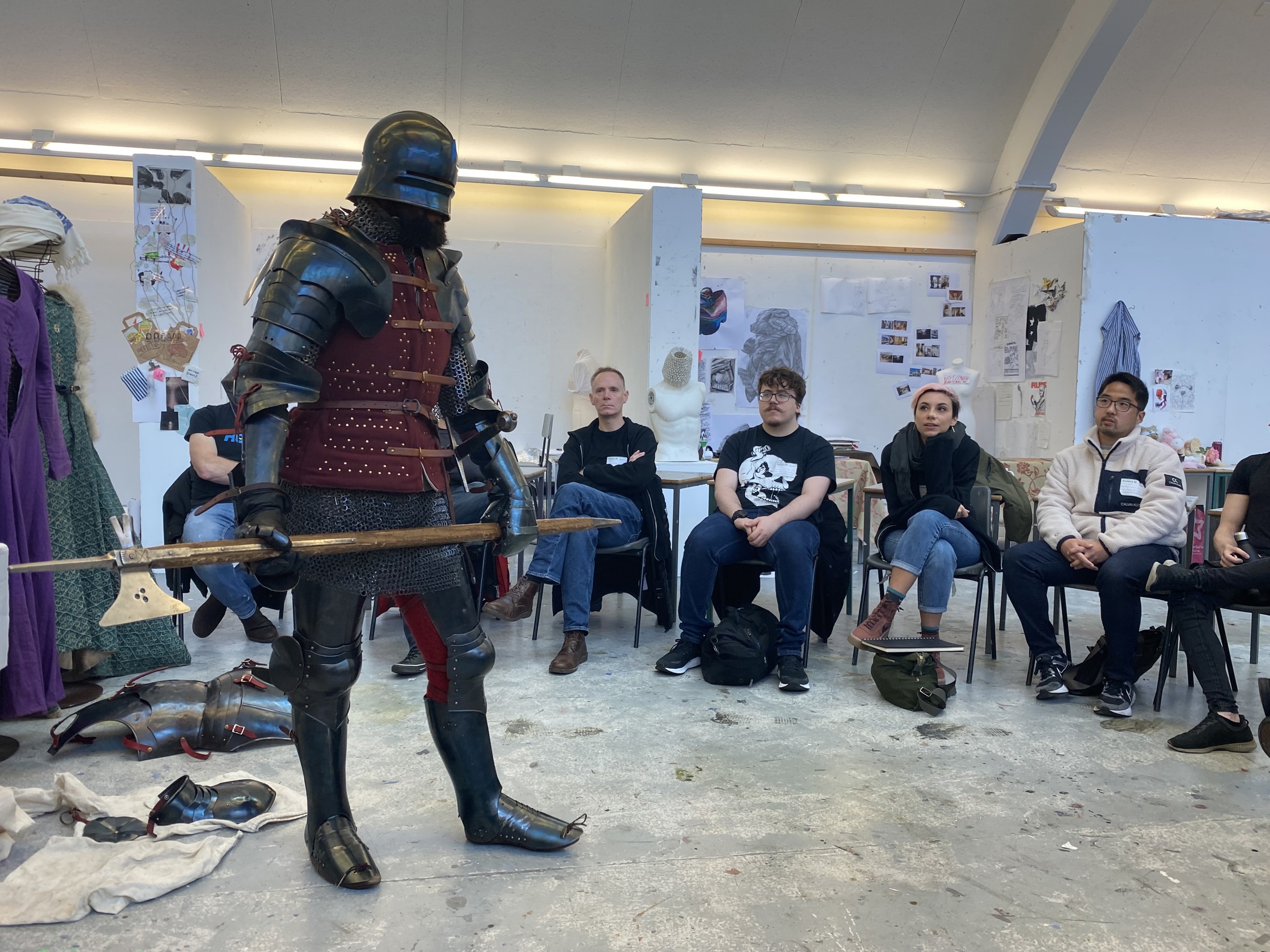 A person wearing medieval armour standing in the middle of an art studio, surrounded by artists